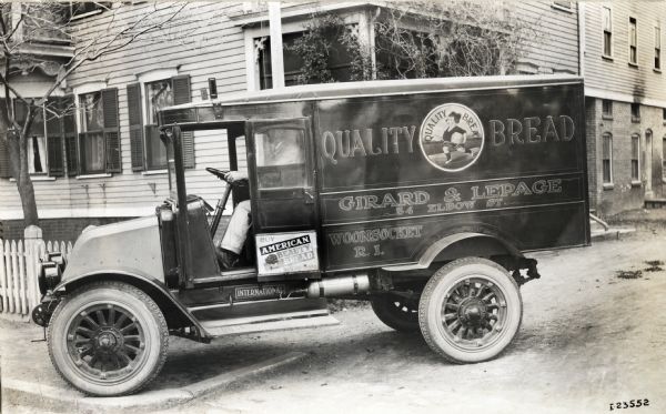 International model "F" or "31" truck operated by Girard and LePage of Woonsocket, Rhode Island, makers of "Quality Bread." A man is sitting in the truck, which is parked in front of a wood frame building. An advertisement on side of the truck reads "Buy American Beauty Bread."