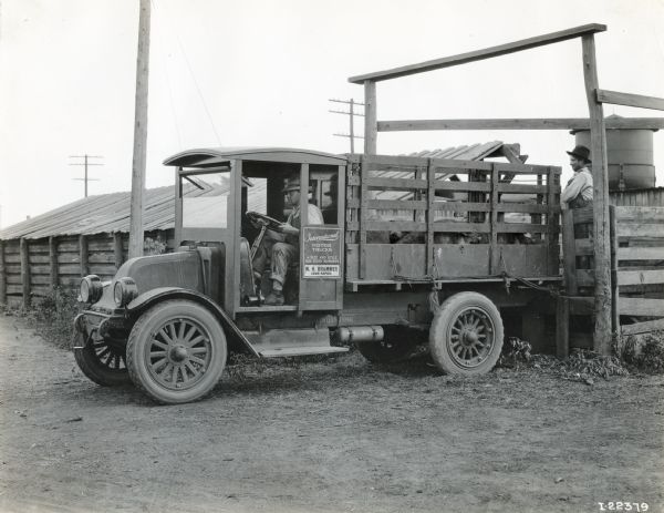 International model "F" or "31" truck operated by W.H. Brammer of Coon Rapids.  One man sits in the driver's seat and another man stands at the back of the truck. The advertising on the side of the truck reads: "International Motor Trucks A Size and Style For Every Business". The truck contains livestock pigs(?) and is photographed in front of buildings and a small water(?) tower.