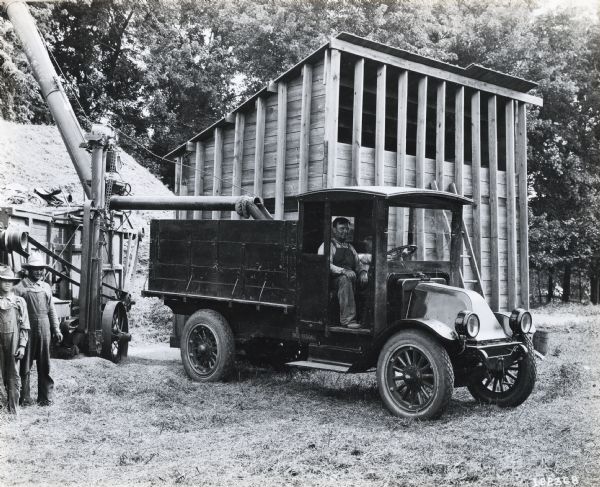 International model "F" or "31" truck. A man is in the passenger seat and a boy sits in the driver's seat. Another man and boy stand beside the truck, which is parked in front of a tall wooden structure.  Agricultural machinery is behind the truck.
