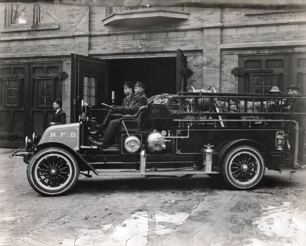 International model "F" or "31" truck operated by N.F.D. (Norwalk Fire Department in Oregon(?)). Four men are in the truck: a driver, passenger, and two men standing in the back. The truck is parked outdoors in front of the firehouse (?) and another man is standing near the garage door.