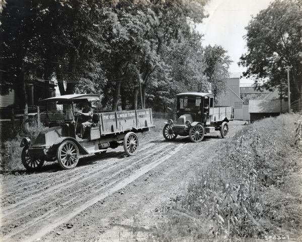 Two International model "F" or "31" trucks operated for Geo. E. Morse Farms of Ladora. Both trucks are driven by male drivers on a dirt road leading from a farm. A house is on the left side of the road.