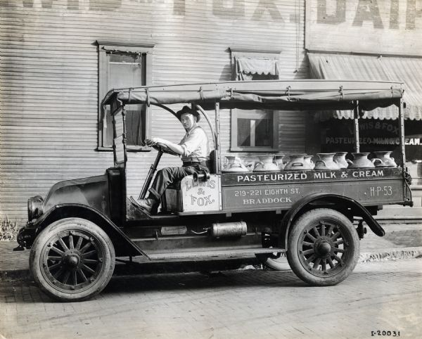 International "F" or "31" truck operated by Lewis and Fox of Braddock (probably Braddock, Pennsylvania). The truck contains milk cans and a man sits in the driver's seat. The truck was photographed on the street outside the Lewis and Fox Dairy. The writing on the truck reads "Lewis and Fox. Pasteurized Milk and Cream 219-221 Eighth St. Braddock."