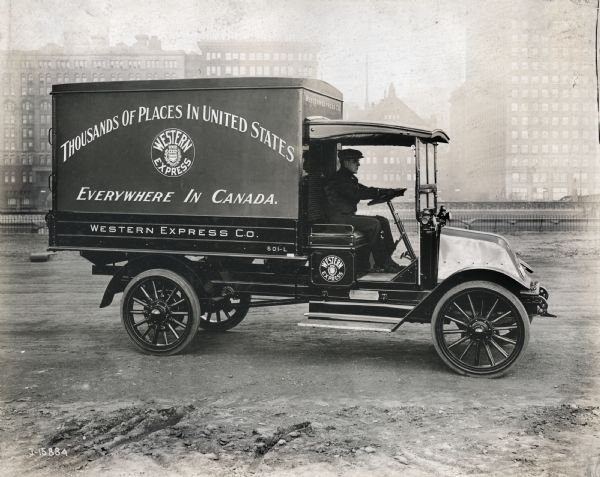 International model "F" truck operated by Western Express Co. There is a man in the driver's seat, and a city skyline in the background.  The advertising on the side of the truck reads: "Thousands Of Places In The United States Everywhere In Canada".