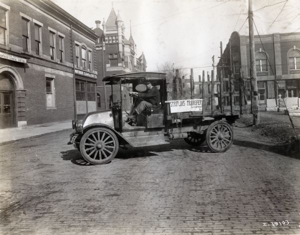 International model "F" "31" truck operated by Striplins Transfer of Litchfield.  A male driver is in the truck which is parked on a city street between a cemetery and the Montrose Cloak and Suit Co. Litchfield Factory.