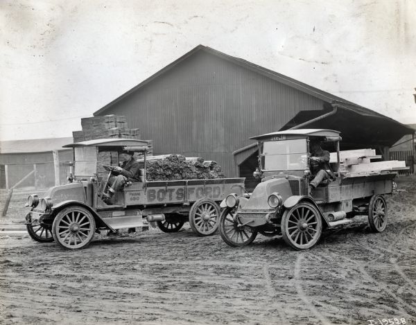 Two International model "F" or "31" trucks operated by Botsford.  Both trucks have male drivers and contain lumber.  The side of the trucks give the Botsford phone number: "Phone 690". In the background is a metal building possibly used for storage.