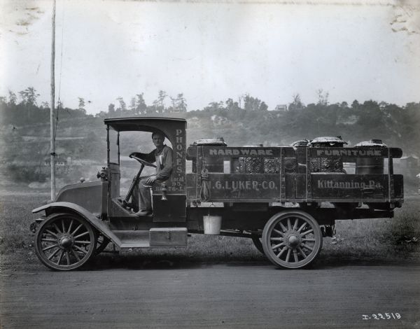 International model "F" or "31" truck operated by H.G. Luker Co. of Kittanning. A man in work clothes is sitting in the driver's seat, and a ridge with homes is in the background. The truck is carrying stoves (?) and has writing on the side of the truck that reads: "Phones Local 95, Be? 76-J" and "Hardware, Furniture." A bucket and rope is also hanging from the side of the truck.