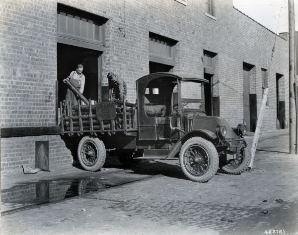 International model "F" or "31" truck operated by Crane Co. in Iowa. The truck is parked outside of a brick building (a warehouse or factory (?)). Two men are loading pipes into the back of the truck from a loading dock.