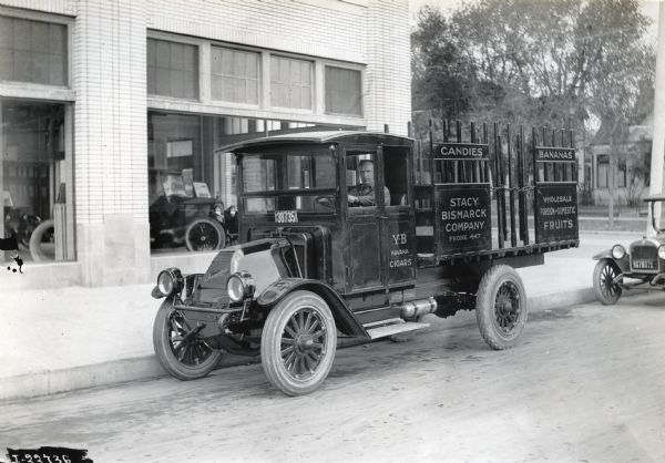 International model "F" or "31" truck operated by Stacy Bismarck Company. The truck is parked on a city street, with possibly a car dealership in the background. A man is seated in the driver's seat of the truck and an advertisment on the side reads: "Candies, Bananas, Wholesale foreign and domestic fruits, YB Havana Cigars" and provides the company's phone number: "Phone 447".