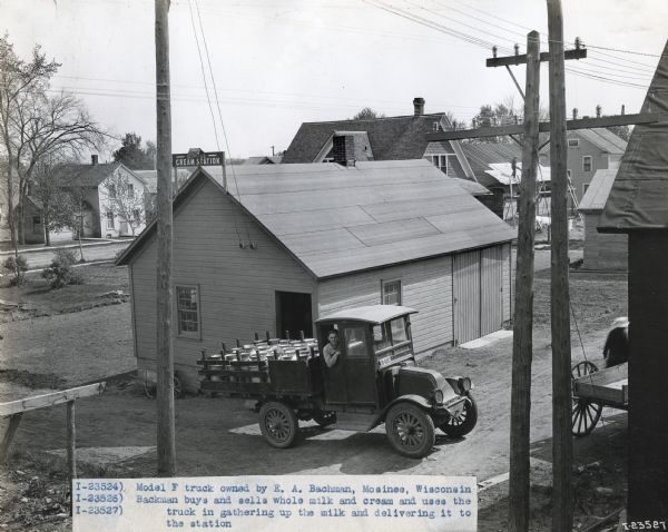 Elevated view of an International model "F" truck carrying milk canisters. There is a man looking out the window of the passenger side of the truck, which is parked outside of a Cream Station. Numerous other wooden buildings are in the background. Original caption reads: "Model F truck owned by E.A. Bachman, Mosinee, Wisconsin. Bachman buys and sells whole milk and cream and uses the truck in gathering up the milk and delivering it to the station."