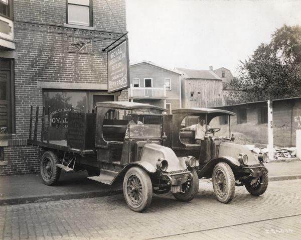 Two International model "F" or "31" trucks with male drivers, which are operated by the Royal Carbonating Company of Providence, Rhode Island. The trucks are parked outside of the Royal Carbonating Co. Advertising on the side of the trucks reads: "Royal Carbonating Co. Royal Club, Ginger Ale, Orange Squeeze Providence, R.I." The company's storefront and sign are in the background along with other buildings.