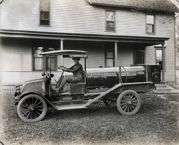 International model "F" truck operated by Standard Oil Co. A man in work clothes sits in the driver's seat. The side of the truck advertises Polarine ("Oil and Greases For Motor Cars") and Burn Perfection Oil. The truck is photographed on the grass in front of a house.
