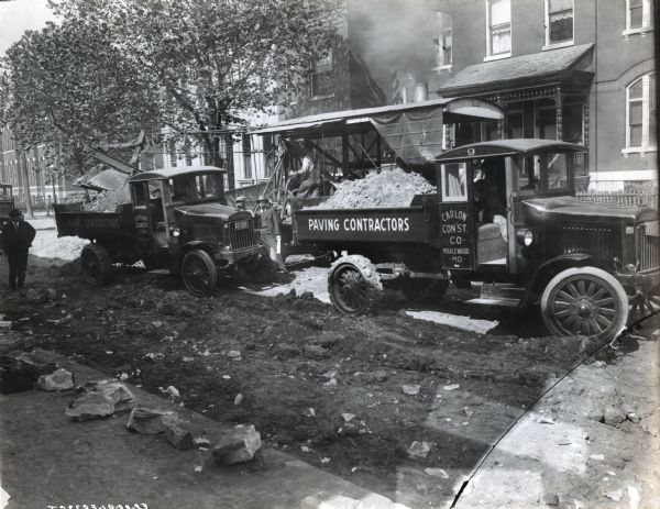 Two International Model 63 trucks operated by Carlon Construction Co. of Maplewood, Missouri. Printing on the side of the truck says "Paving Contractors." The trucks were photographed carrying rocks and sediment, in a road construction area, with homes in the background. Several men also appear in the photograph; one driving a truck, three standing beside the truck (one carrying plans(?)),and one operating equipment.