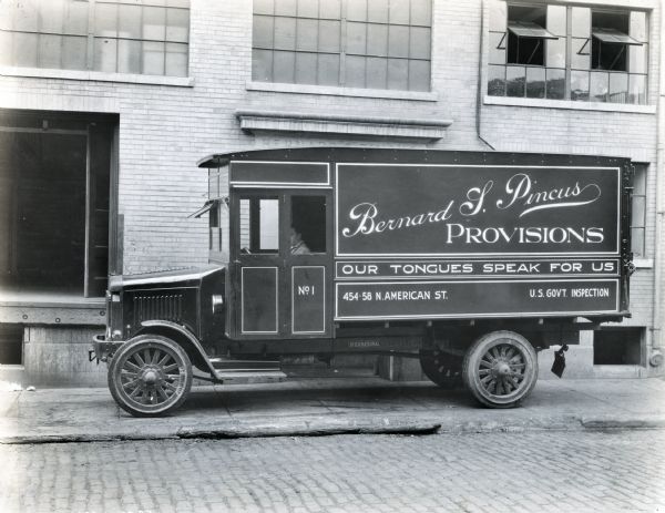 International Model 63 truck operated by Bernard L. Pincus Provisions.  The truck was photographed with a man sitting in the driver's seat outside a brick building with a loading dock. Printed on the side of the truck is the slogan, "Our Tongues Speak For Us," "454-58 N. American St.," and "U.S. Gov't. Inspection."
