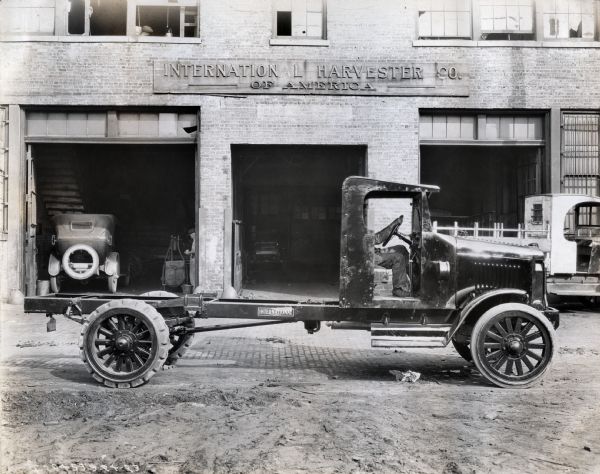 International Model 63 Truck.  Truck was photographed outside the International Harvester Co. with a man seated in the driver's seat. Three garages are pictured in the background and several other vehicles are also pictured.