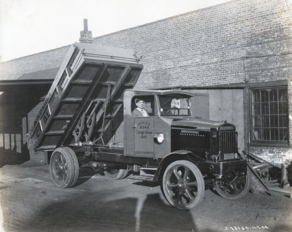 International Model 63 truck operated by Arquilla Bros. There is a man sitting in the passenger's seat outside of a brick building (possibly the coal storehouse). Printed on the side of the truck is "Office 9348 Cottage Grove Ave." Printed below the grill of the truck is "The Coal Number" "Chesterfield O111".  The back of the truck is elevated as if it were dumping a load of coal.