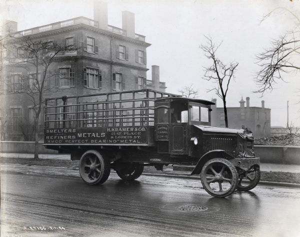 International Model 63 truck operated by H. Kramer and Co. The truck is on the street outside a large house(?) with a man seated in the cab.  Printed on the side of the truck is the company name, address, phone number, and "Metals" "Smelters and Refiners" "'KMCO' Perfect Bearing- Metal".