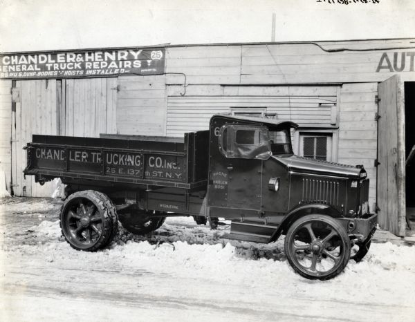 International Model 63 truck operated by Chandler Trucking Co. of New York City. The truck is parked in the snow outside the Chandler and Henry general Truck Repairs building which advertises "Repairs of U.S. Dump-Bodies" "Hoists Installed." Printing on the side of the truck reads, "25 E. 137th St. N.Y." "Phone Harlem 8051."

