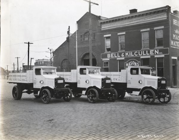 Three International Model 63 trucks operated by Bell and Kilcullen Inc. of New York. The three trucks are parked outside the Bell and Kilcullen store which advertises itself as a Mason shop. The company's phone number is printed on the side of the trucks and the trucks are labeled 1, 2, and 3.