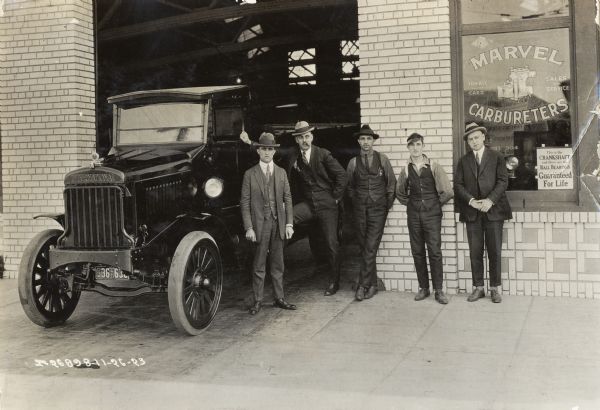 International Model 63 truck photographed outside a store or garage (possibly an International dealership). A shop window advertises Marvel Carbureters. The truck is parked at the entrance to the garage and five men are standing next to it.