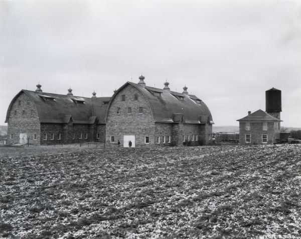 Two men stand in the distance outside one of the farm buildings near a field at St. Marvis farm, where a McCormick-Deering milk cooler is installed. There is a silo or water tower in the background.