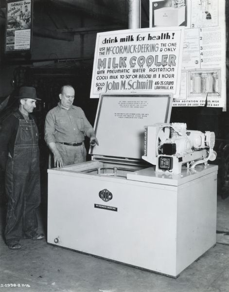 John M. Schmitt (probably an International Harvester dealer), right, shows a customer a McCormick-Deering 6-can milk cooler on display. The sign above the cooler reads: "Drink Milk for Health! Use the McCormick-Deering; The One & Only Milk Cooler with Pneumatic Water Agitation Cools Milk to 50 degrees or Below in 1 Hour; Sold by John M. Schmitt." The display may be at Mr. Schmitt's dealership, or at a fair or other local event.