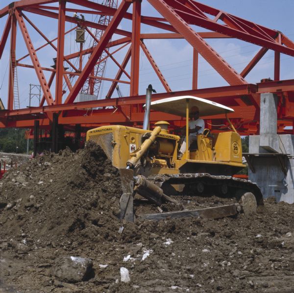 Color photograph of a worker operating an International TD-20C crawler tractor (TracTracTor) at a bridge construction site. Other construction workers are on the bridge's support structure. The construction firm was Bates and Rogers Contracting Corp.