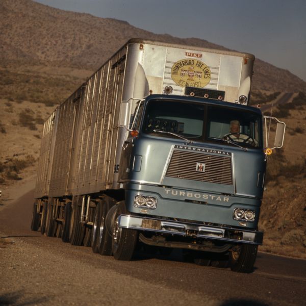 Color photograph of a man driving an International Turbostar truck on a highway in a mountainous landscape. The location is likely International Harvester's Phoenix Proving Ground.