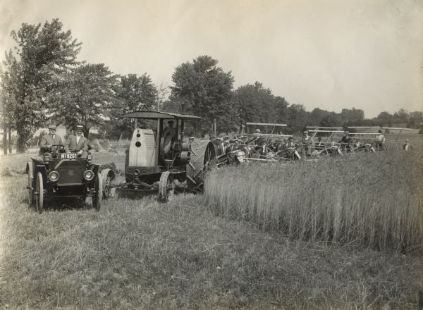 A large group of men standing in a field where two men in an International Auto Wagon lead a procession of an International tractor and several grain binders(?).