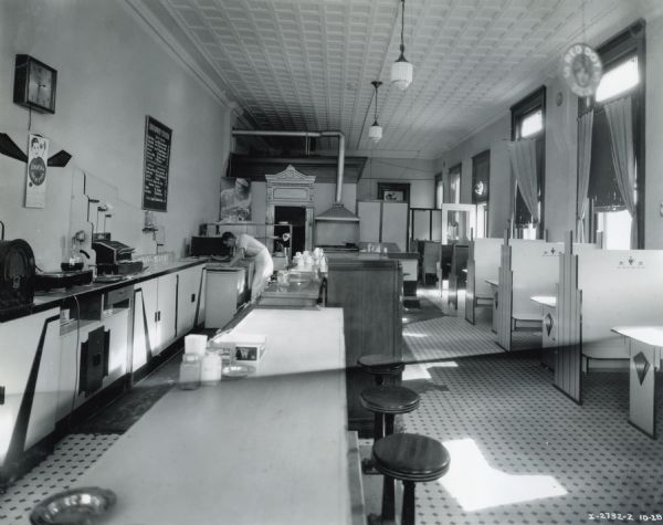 Interior of Reeds Cafe in Elsberry, Missouri. There is a man at the cafe reaching into a 6-can milk cooler.  Also shown is the work space behind the counter including a radio, coffee machine, cash register, glasses, a menu, a clock, and an advertisment for "Joe Bonansinga and His Orchestra" "At Elsberry Mo Fri Oct 30 Halloween Dance".  There is also a lunch counter and booths.
