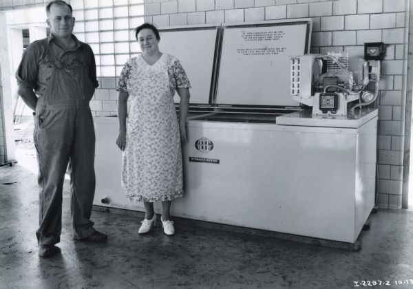 Mr. and Mrs. John Kalbrach stand in front of their McCormick-Deering 10-Can Milk Cooler. Mr. Kalbrach is wearing overalls and Mrs. Kalbrach is wearing a flowered dress and white lace-up high-heel shoes.