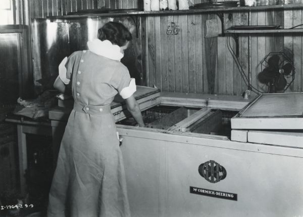 A woman wearing a dress places bottles into a "McCormick-Deering Milk Cooler "for cooling beer". The photograph was taken at Royce Cafe in Edmond, Oklahoma. Also seen in photograph are the wooden paneled walls and shelves.