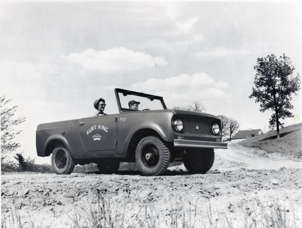 A man and a woman are sitting in a four-wheel drive, open-top International Harvester Scout.  The text on the side of the truck reads: "Kurt King." The attached press release reads: "The new all-purpose Scout, an International Harvester Company product, can be converted to an open runabout by easy removal of its steel passenger-compartment top. Also removable are the Scout's door glass and doors.  Its front windshield folds down. The low-cost Scout has a five-foot-long integral pickup body with extended wheel housings which can serve as additional seating. It is available in a two-wheel drive model and a four-wheel drive model (above).  Power for both models is provided by a new International Comanche four-cylinder engine rated at approximately 90 horsepower. Wheelbase and overall length for both are 100 inches and 12 feet, ten inches, respectively. Scout suggested retail prices begin at $1598, plus federal and local taxes, FOB, Fort Wayne, Ind."