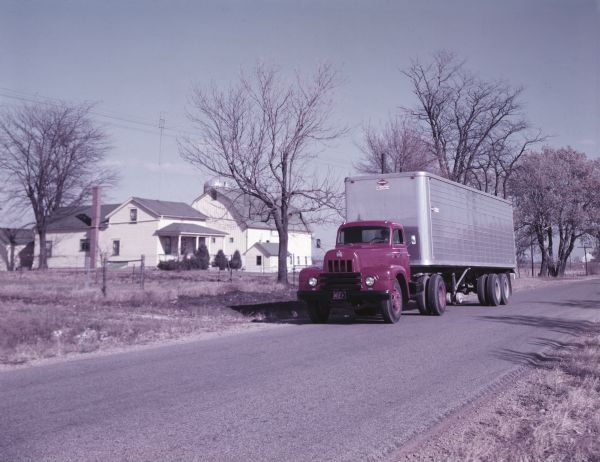 Color photograph of a man in a uniform sitting in an International Harvester R-195 semi-truck outfitted with a Space Saver cab. The truck is parked along a road near a farm.