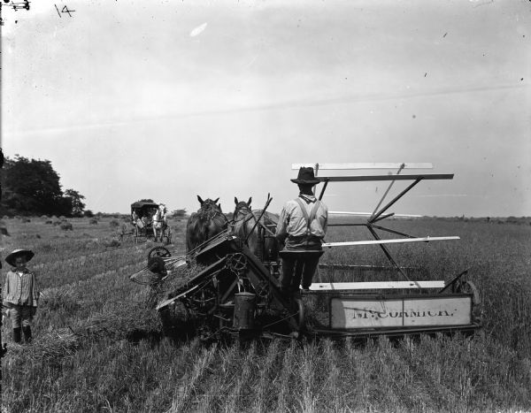 A farmer is operating a McCormick grain binder pulled by two horses in a field. A young child wearing a wide-brimmed hat is watching, and a horse-drawn buggy carrying several people is approaching the farmer from a distance.