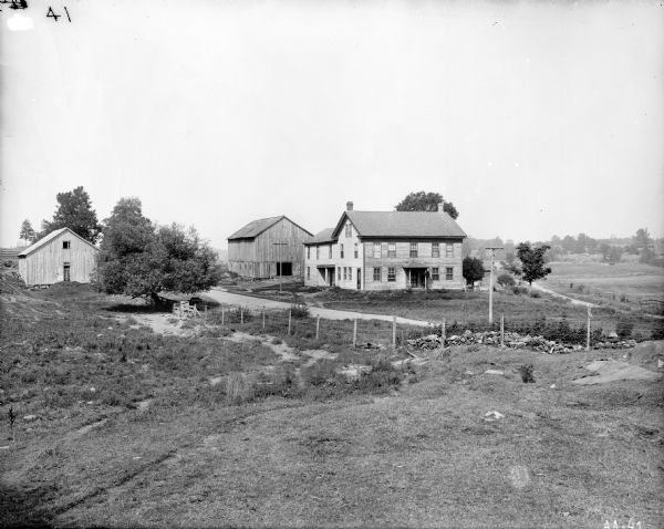 View over field of two-story frame farmhouse with wooden barn to the rear and a smaller shed off to the side. A barbed wire(?) fence and a utility pole are in the foreground.