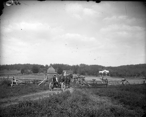 A farmer is operating a mower pulled by two horses towards a farmstead. A farmhouse, barn, and outbuildings, one with a cupola, are in the distance.