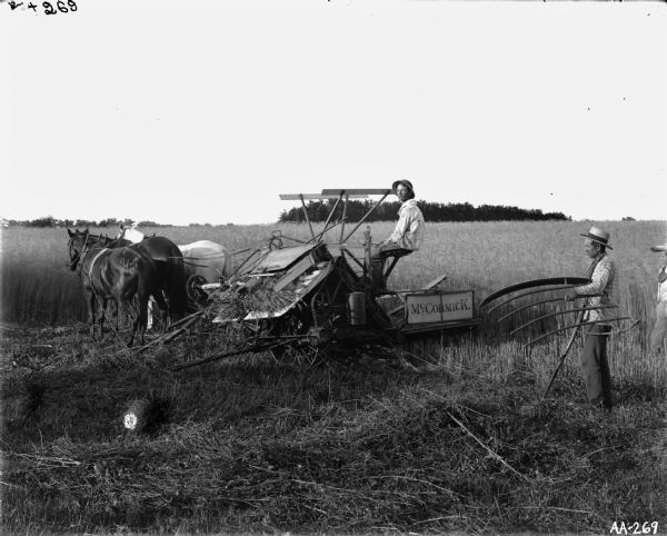 Three farmers harvest grain with McCormick horse-drawn grain binder. One farmer is sitting aboard the binder guiding horses while two men, one with scythe and cradle, are standing behind waiting for bundles.