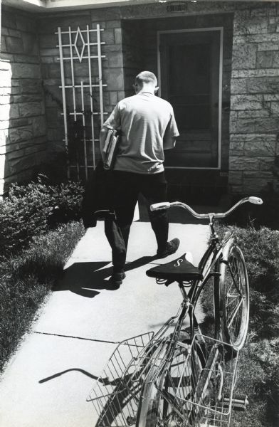 Thirteen-year-old John "Butch" Quick is photographed returning to his house. Butch carries his jacket and books under his arm as he enters his house. The foreground captures a (?Schwinn) bicycle with two carrier baskets. Original magazine caption reads: "School out for the day, bike wheels give way to Cub Cadet wheels for Butch Quirk. Books are dropped off, clothes changed, bucket of seed grabbed up."