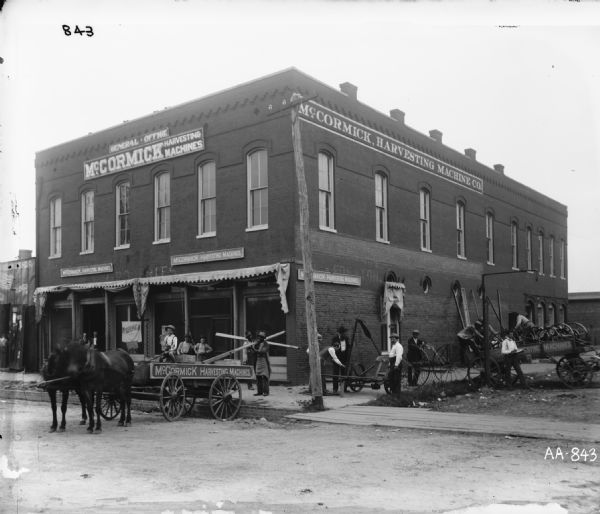 View from street of workers, including some African Americans, loading (and/or unloading) agricultural equipment and parts outside a general office of the McCormick Harvesting Machine Company. The building was located at Lafayette and Shannon. The men pose with horse-drawn wagons, a mower and a hay rake.