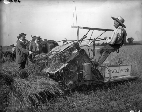 Three men, one on binder, and two standing, are posing with a horse-drawn McCormick grain binder in field as farmhands are bundling grain in the background.