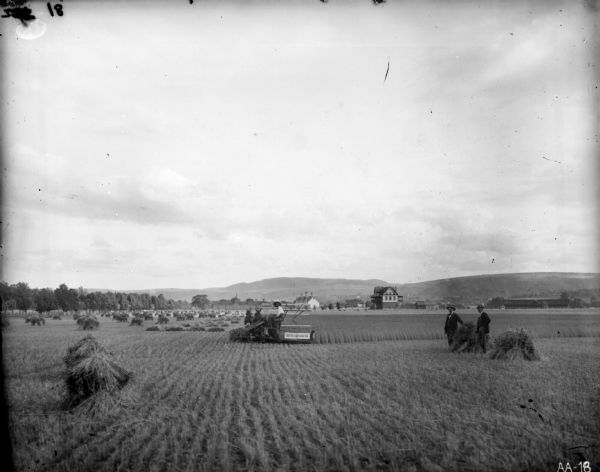 View from a distance of a farmer posing on a McCormick grain binder drawn by two horses in a wheat field. Two men in suits are posing next to a bundle of wheat to the right of the farmer, and numerous bundles of wheat are in the field to his left. Farm buildings are in the background.