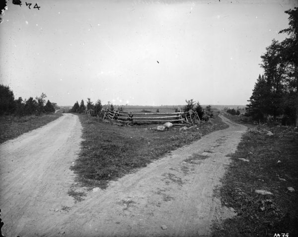 View of fork in road, with split-rail fence in the center. There are buildings in the far background on the left.