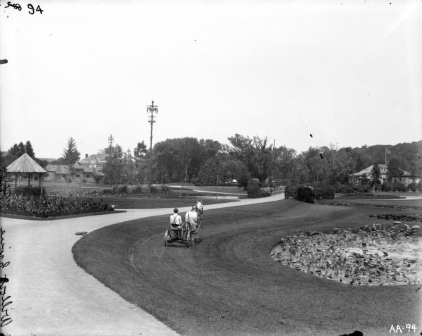 Elevated view of a male grounds keeper operating a horse-drawn mower on the grounds of an estate. A pond with lily pads and a gazebo are in the foreground, and a house is in the distance. There are also several electric street lamps.