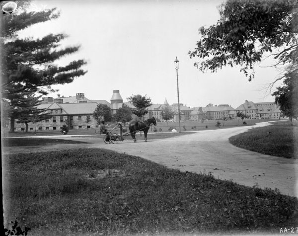 A man is driving a horse-drawn mower down a path on the grounds of a large estate. There is an electric street lamp along the path, and many buildings in the background.
