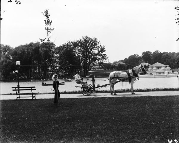A male grounds keeper is driving a horse-drawn mower down a path on a park or estate grounds. Another man is leaning against a slender tree. Both men are wearing hats, white shirts and suspenders. There is a body of water in the background, perhaps a pond or stream, on which several people are canoeing. There is also a fountain, several electric street lamps, and a pavilion on the water.
