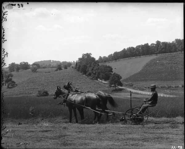 A man sporting a beard and wide-brimmed straw (?) hat is operating a mower pulled by two horses in a field. The field is on hilly terrain. A dirt road and split-rail fence are in the distance. "E Rees Howard Farm. Newton Square Phila" is etched into the emulsion of the image.