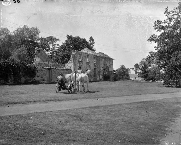 A farmer operating a horse-drawn mower on the grounds in front of a brick building in ruins.