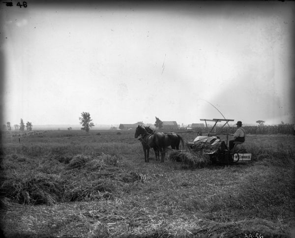 A farmer wearing a bowler or derby hat is voperating a horse-drawn McCormick grain binder in a field. The farmer is holding a switch. A cornfield and several houses and farm buildings can be seen in the distance.