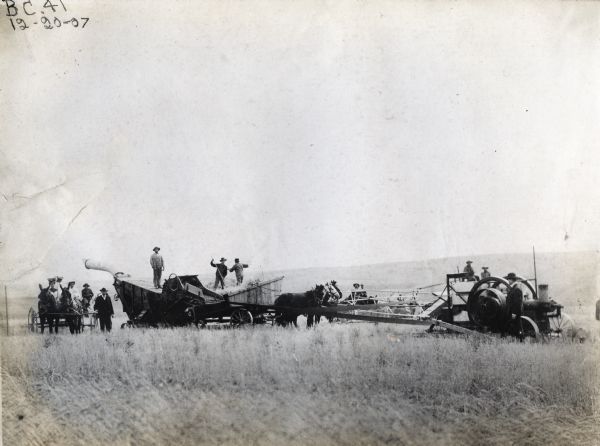 Men are working with horses, a threshing machine, a push binder(?) and stationary engine in a field. A group of men and women with a horse-drawn buggy are looking on from one side.
