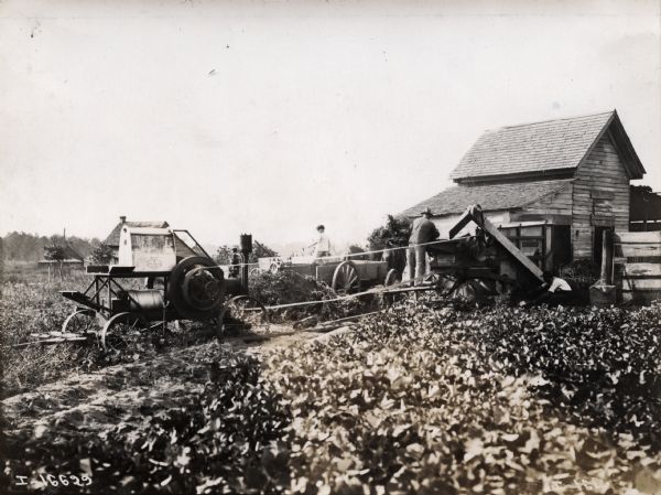 Men are operating a thresher and a stationary engine near a barn.